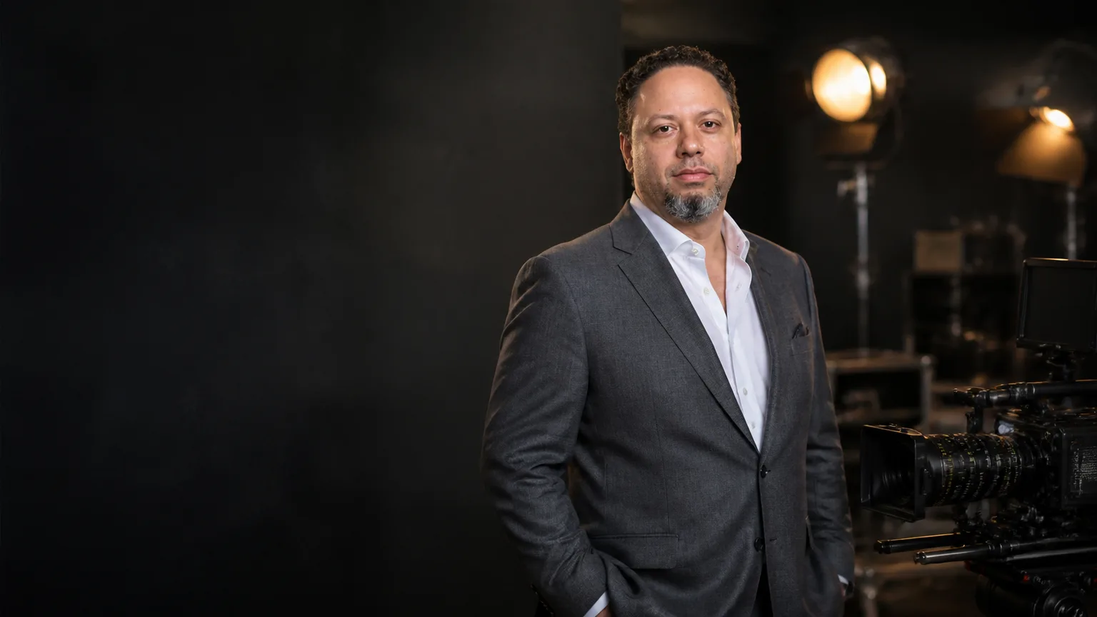 Tristan Desechenes in a tailored gray suit beside a cinema camera on a softly lit soundstage